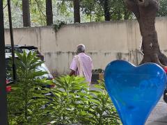 An old man walking on the street, as seen through a cafe window. Wearing a pink shirt, white lungi, and a towel on his shoulder.