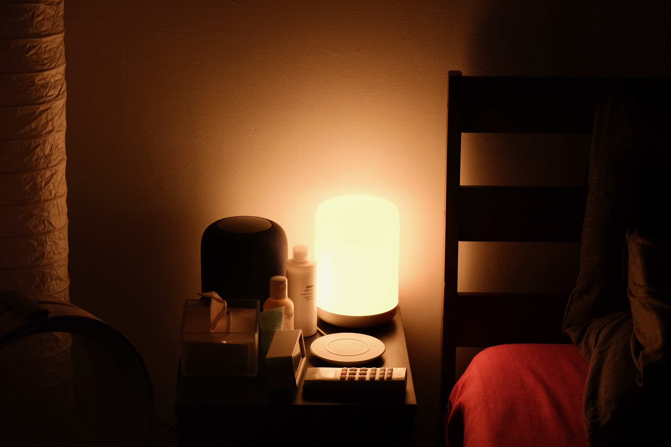 A bedside table with a warm light source, a speaker, some bottles of lotion, tissues, a clock, a remote, and a white wireless charger.