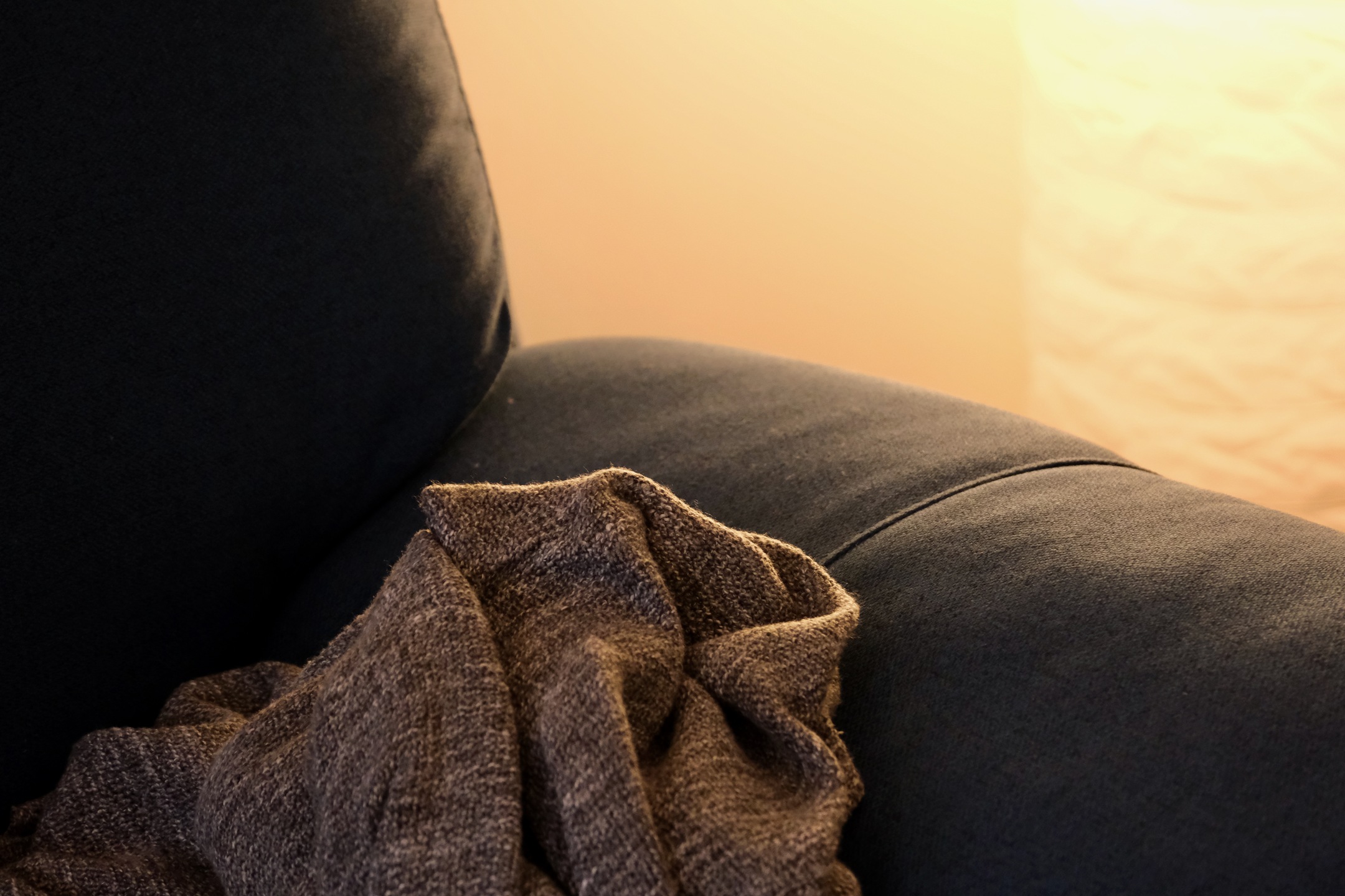 A close up shot of a dark blue fabric couch with a gray throw blanket. There is a warm source of light behind the couch.
